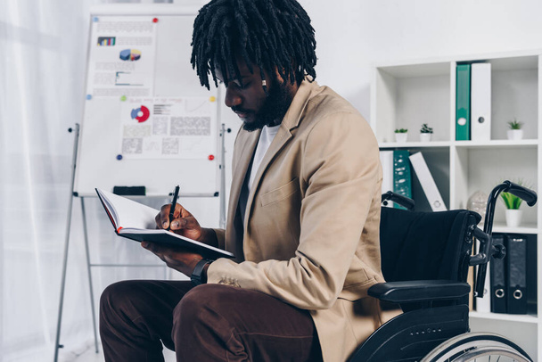 African american disabled employee on wheelchair writing in notebook in office - Foto, immagini