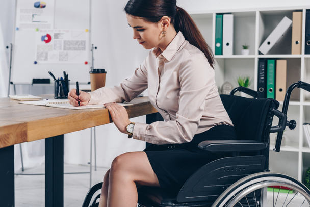 Disabled employee on wheelchair writing in notebook at table in office - Fotografie, Obrázek