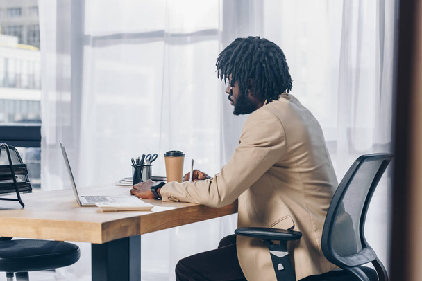 Selective focus of african american recruiter with laptop and paper cup of coffee at table in office - 写真・画像