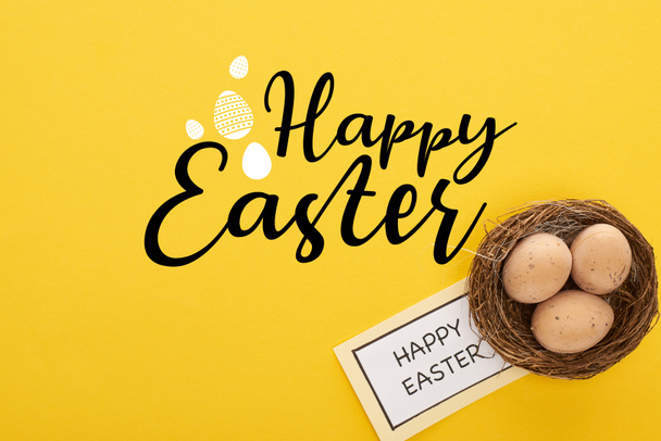 top view of greeting card with happy Easter lettering near chicken eggs in nest on yellow colorful background - Fotografie, Obrázek