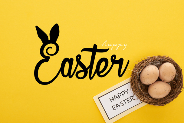top view of greeting card with happy Easter lettering near chicken eggs in nest on yellow colorful background - Photo, Image
