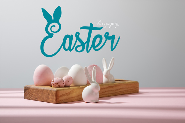 Πασχαλινά αυγά σε ξύλινη σανίδα με διακοσμητικά κουνέλια σε ροζ και γκρι με χαρούμενη πασχαλινή απεικόνιση - Φωτογραφία, εικόνα