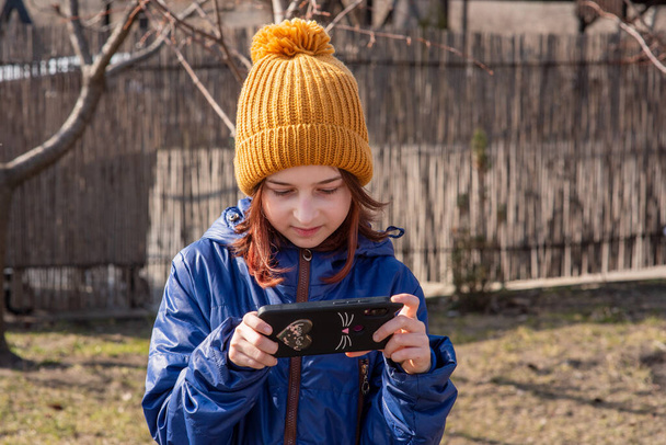 Girl dressed in jacket, poses for making selfie or photo of herself with smartphone. People and leisure concept. Girl makes selfie in a red jacket.Girl 9 years old with a smartphone in her hands. Teen - Photo, Image