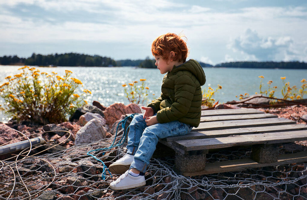 lindo pelirrojo niño sentado en paleta de madera en frente del mar Báltico y flores de tanaceto
 - Foto, imagen