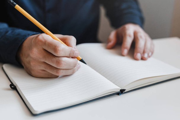 Closeup of the man's hands holding a pencil between fingers and writing in a notepad. Notebook on the table. Young businessman working in an office. Business, finance, industry and job concept - Photo, Image