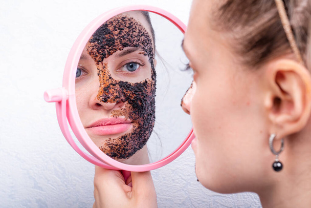 Młoda atrakcyjna kobieta nakłada fartuch na twarz, patrząc w lustro - peeling do mycia kawy - Zdjęcie, obraz