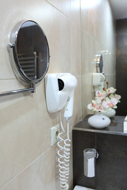 A hair dryer hangs on the wall . Photo in the interior. Flowers and a mirror are out of focus. The concept of the hotel business. Side view. Copy space. - Photo, Image