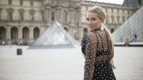 Beautiful blond woman in transparent dotted dress walking in front of the Louvre museum on a sunny summer day. Action. Young female smiling and walking near the glass pyramids. - Fotó, kép