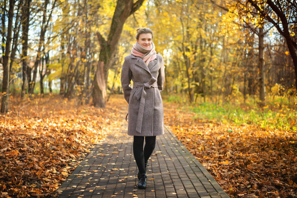 Nuori viehättävä nainen takissa kävelee syksyllä puistossa polkua pitkin langenneiden lehtien kanssa.
 - Valokuva, kuva