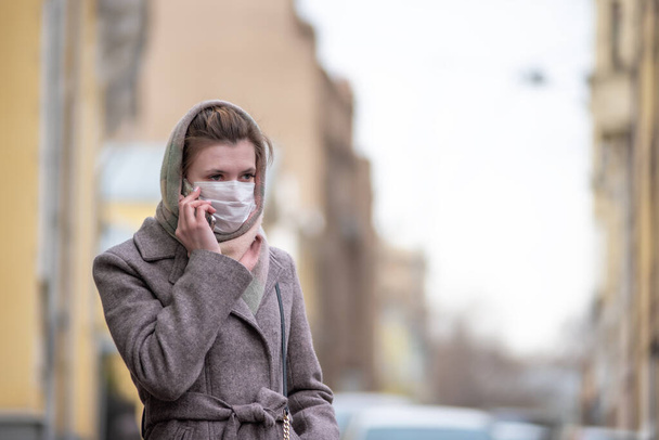 Young attractive European Caucasian woman in a medical protective mask against influenza viruses and coronavirus COVID-19 talking on the phone on a city street - Φωτογραφία, εικόνα
