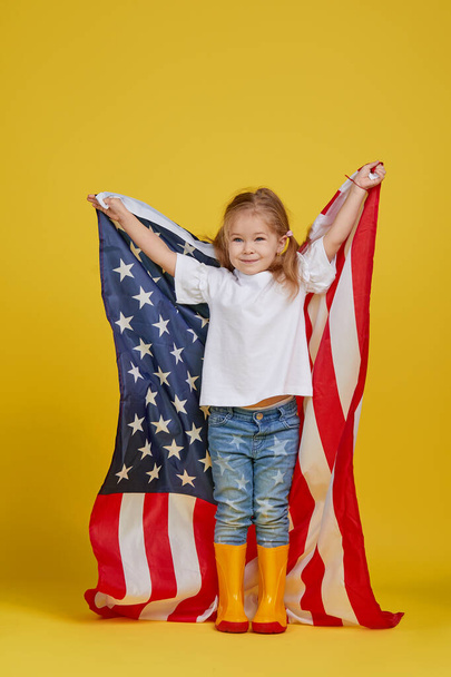 Patriotic holiday. Happy child, cute little girl with american flag on yellow studio background. USA celebrate July 4th, independence Day. - Foto, imagen