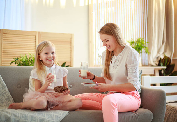 Young mother with child daughter at home on the couch drinking milk and cookies, laughing and talking together - Photo, Image
