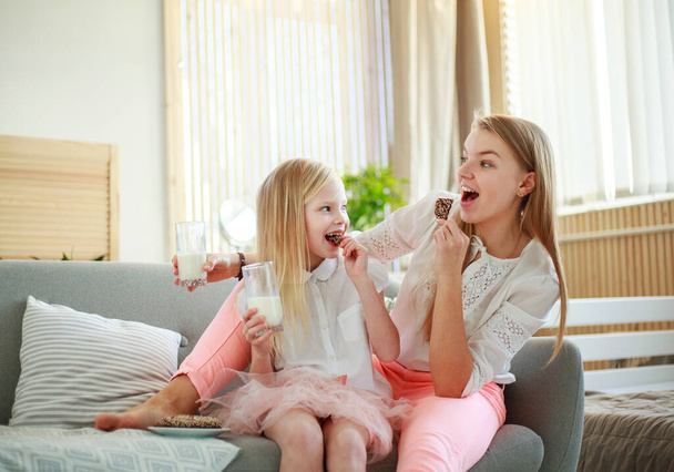 Young mother with child daughter at home on the couch drinking milk and cookies, laughing and talking together - Photo, image