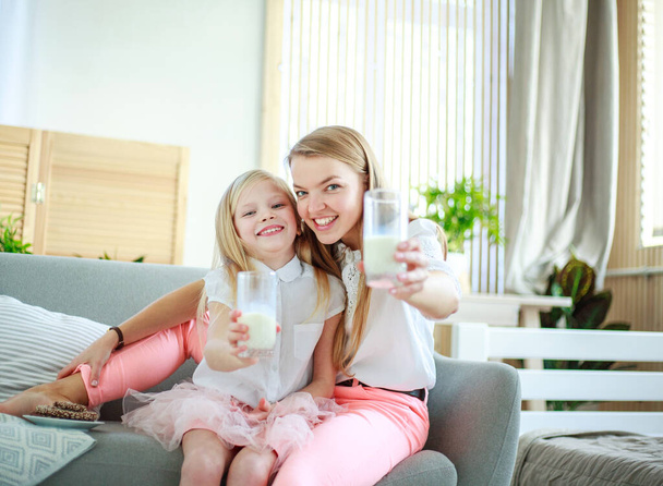 Young mother with child daughter at home on the couch drinking milk and cookies, laughing and talking together - Foto, imagen