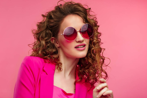 Close up fashion studio portrait of young beautiful women with curly hair.Nice girl with wavy hairstyle,bright make up,stylish eyeglasses isolated over pink background. Beauty and hair care concept - Photo, image