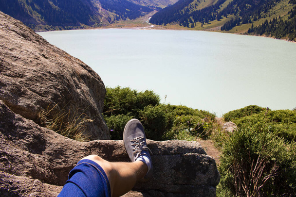 Ноги отдыхают в горах рядом с спокойным озером воды, травы и лодки. Туристическая концепция
 - Фото, изображение