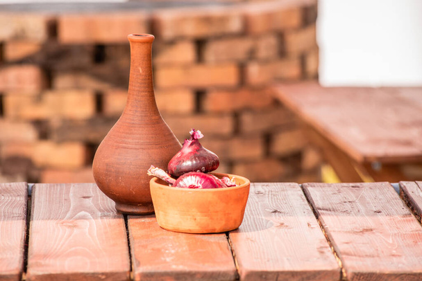Garrafa de barro e tigela com cebolas vermelhas em uma mesa de madeira
 - Foto, Imagem