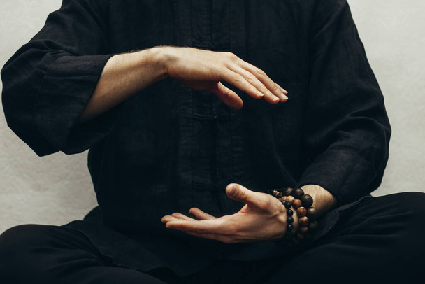 Facet w czarnej koszuli siedzi i robi qigong. Ręce prosto na energię. Modlitwa, wdzięczność. Praktykujący mnich. Energia Qi. Stanowisko do jogi. Zamknij się. - Zdjęcie, obraz