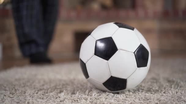 Крупним планом футбольний м'яч лежить на підлозі в приміщенні. Чоловічі ноги в піжамі б'ються і слідують за нею. Концепція здорового способу життя, спортивна активність, життєва важливість
. - Кадри, відео