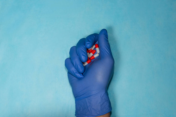 Крупним планом рука лікаря в синіх медичних рукавичках стискає червоні та білі таблетки
 - Фото, зображення