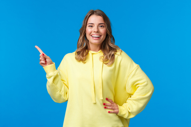 Take a look. Upbeat good-looking blond girl pointing finger left, recommend promo, advertise product, look pleased and smiling camera, explain project, standing blue background - Photo, Image
