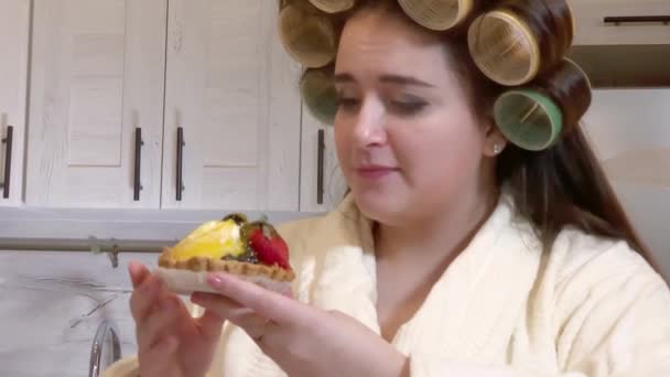 Plus-size girl, dressed in a bathrobe, curlers on her head, looks at the cake with desire, not daring to start eating it - Video, Çekim
