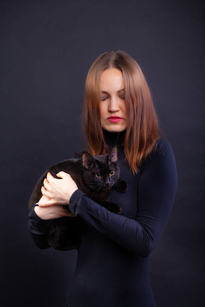 red-haired young woman in black clothes on a black background with a black cat - Foto, Imagen