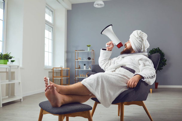 Funny pedicure beauty mask cosmetology concept. Funny fat man in a bathrobe shouts through a megaphone in room salon - Foto, imagen