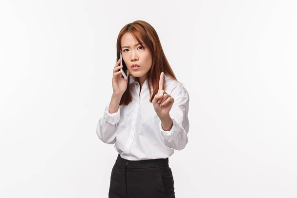 Минутку, минутку. Занятая, серьезная деловая женщина, разговаривающая по телефону, показывающая пальцем и просящая кого-то подождать, пока она закончит говорить, стоять на белом фоне
 - Фото, изображение