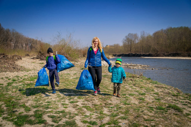 joven rubia cabello mujer con sus hijos en guantes azules limpieza de la orilla del río de plástico y basura en grandes bolsas de basura azul en un día soleado a principios de primavera
 - Foto, imagen