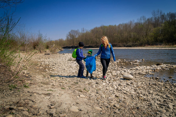 mladá blond vlasy žena se svými syny v modrých rukavicích čištění břehu řeky z plastu a odpadky ve velkých modrých pytlů na odpadky na slunný den na začátku jara - Fotografie, Obrázek