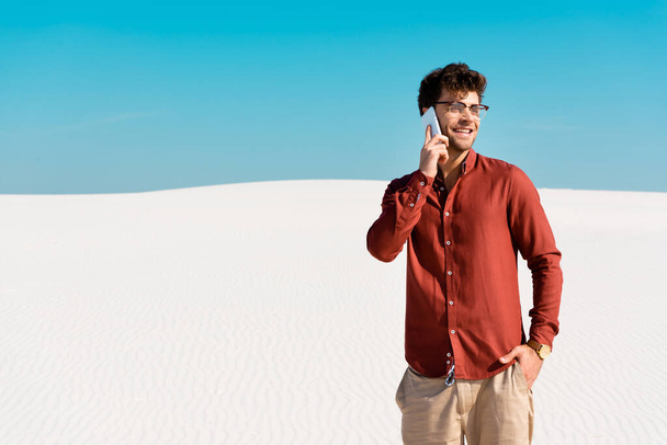 smiling handsome man on sandy beach talking on smartphone against clear blue sky - Foto, afbeelding