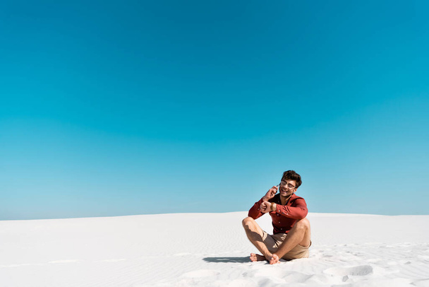 onnellinen mies hiekkarannalla puhuu älypuhelimella ja katselee rannekelloa kirkasta sinistä taivasta vasten
 - Valokuva, kuva