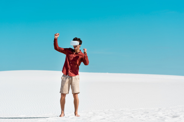 excited handsome man on sandy beach in vr headset against clear blue sky - Foto, imagen