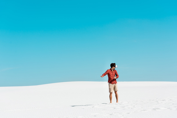 man on sandy beach in vr headset gesturing against clear blue sky - Foto, Imagem