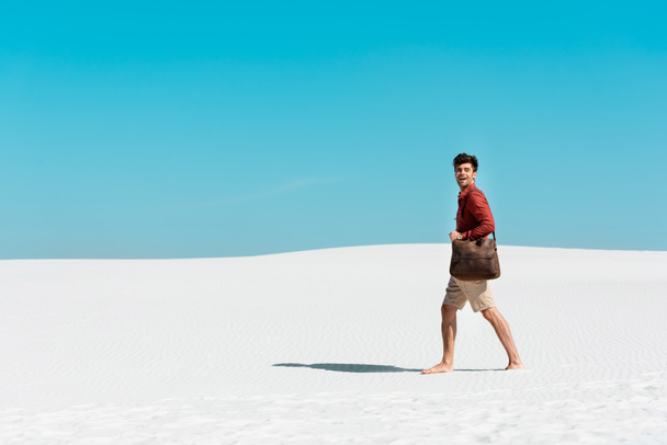 Seitenansicht eines gutaussehenden Mannes mit Ledertasche, der am Sandstrand vor blauem Himmel spaziert - Foto, Bild