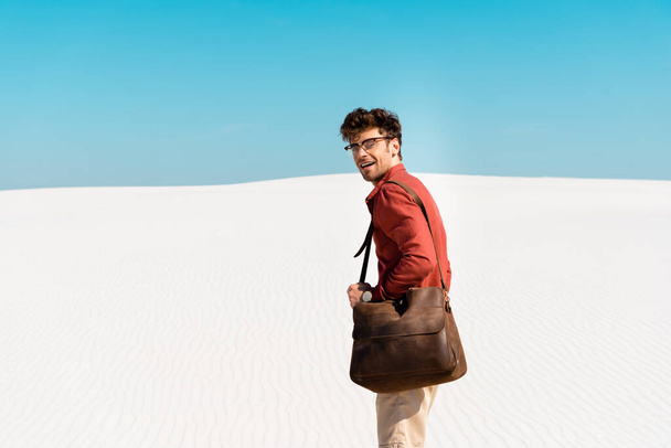 smiling handsome man with leather bag on sandy beach against clear blue sky - Photo, image