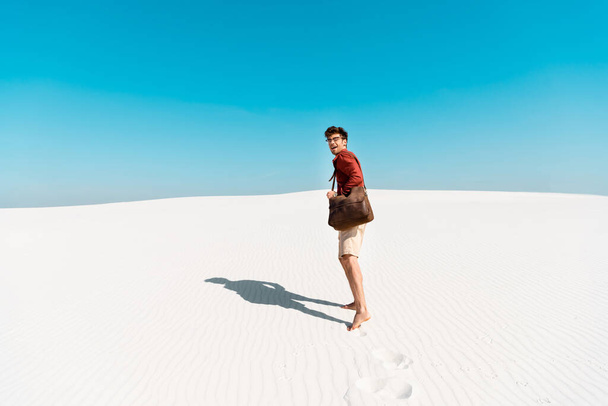όμορφος άντρας με δερμάτινη τσάντα σε αμμώδη παραλία με πατημασιές - Φωτογραφία, εικόνα