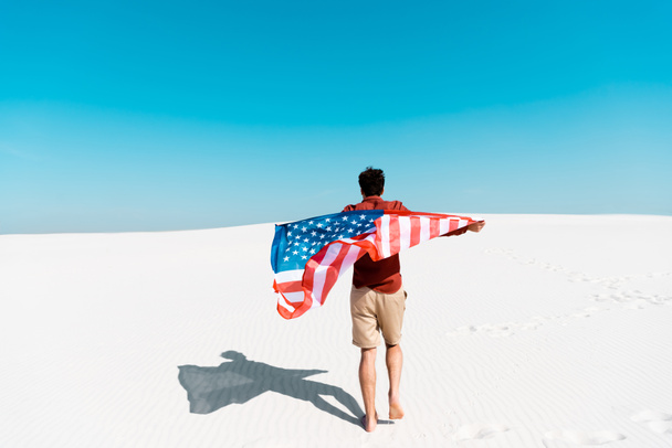 widok z tyłu człowieka z amerykańską flagą na wietrznej piaszczystej plaży z jasnym błękitnym niebem - Zdjęcie, obraz