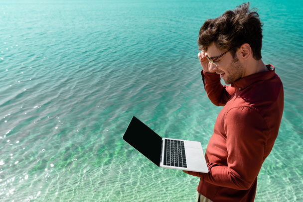 side view of smiling handsome freelancer with laptop standing in turquoise water - 写真・画像