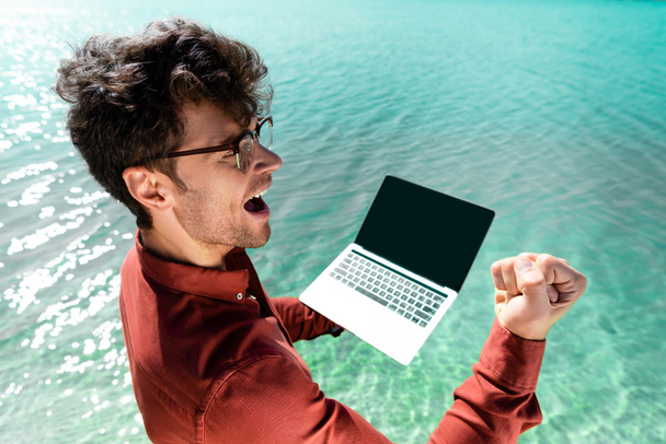excited handsome freelancer with laptop standing in turquoise water - Foto, imagen