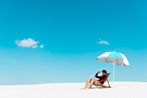 freelancer zittend met laptop in ligstoel onder parasol op zandstrand tegen blauwe lucht - Foto, afbeelding
