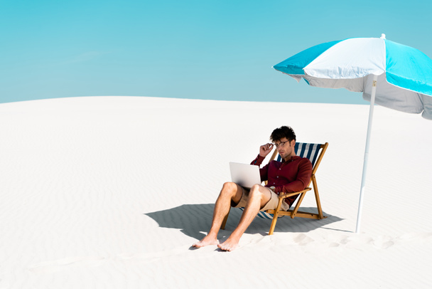 freelancer κάθεται με φορητό υπολογιστή στο κατάστρωμα καρέκλα κάτω από ομπρέλα στην αμμώδη παραλία κατά μπλε ουρανό - Φωτογραφία, εικόνα