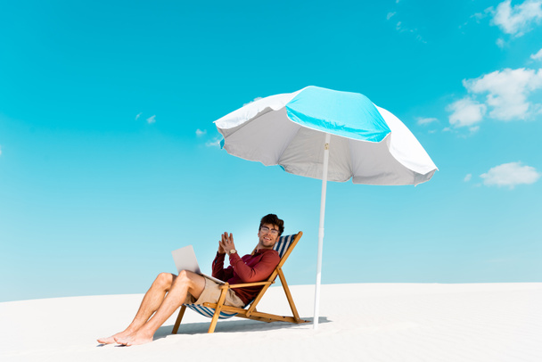 smiling freelancer sitting with laptop in deck chair under umbrella on sandy beach against blue sky - Fotó, kép