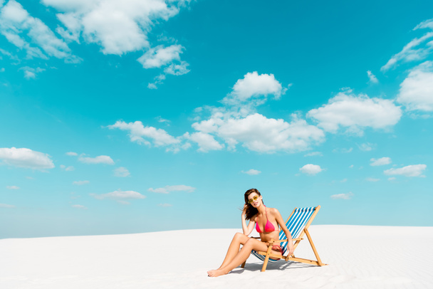 Mayolu ve güneş gözlüklü güzel bir kız kumlu sahilde mavi gökyüzü ve bulutlu bir sandalyede oturuyor. - Fotoğraf, Görsel
