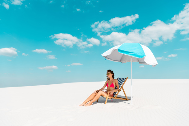 smiling beautiful sexy girl in swimsuit and sunglasses sitting in deck chair under umbrella on sandy beach - 写真・画像