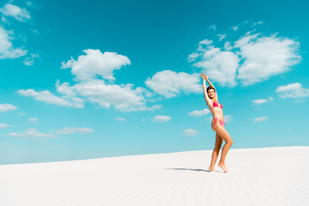 smiling beautiful sexy girl in swimsuit with hands in air on sandy beach with blue sky and clouds - Фото, зображення