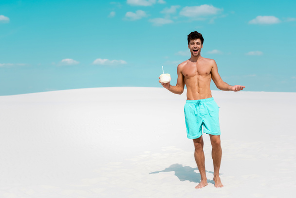 gelukkig sexy man met gespierde romp in zwemmen shorts met kokosnoot drinken op zandstrand - Foto, afbeelding