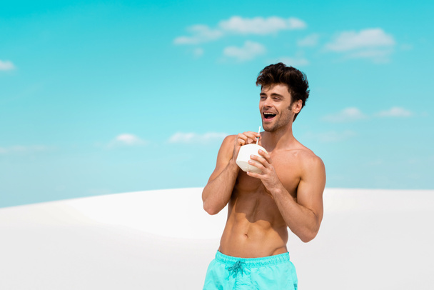 sonriente hombre sexy con torso muscular en pantalones cortos de natación con bebida de coco en la playa de arena
 - Foto, imagen