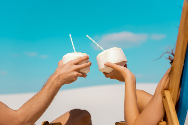 cropped view of young couple sitting in deck chairs with coconut drinks on sandy beach - Photo, Image